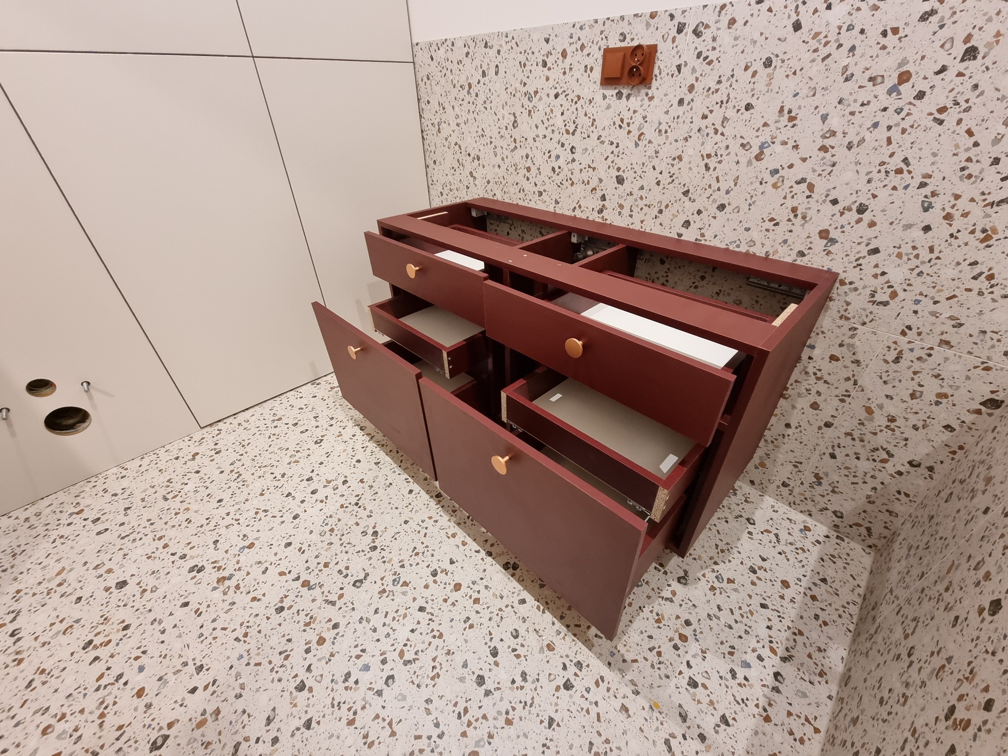 Częściowo zmontowana szafka łazienkowa z otwartymi szufladami w kolorze bordowym, z okrągłymi złotymi uchwytami, na tle ściany i podłogi wykończonej płytkami lastryko.