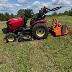 Czerwony traktor Yanmar kosi trawę na polu w słoneczny dzień. Świeżo skoszona trawa leży na ziemi. Widoczny fragment pola i drzewa w tle.