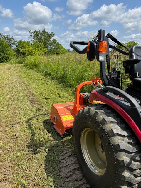 Czerwony traktor z kosiarką rotacyjną na polu, ślad koszenia widoczny w trawie. Błękitne niebo z chmurami w tle. Kąt ujęcia z bliska, z boku maszyny.