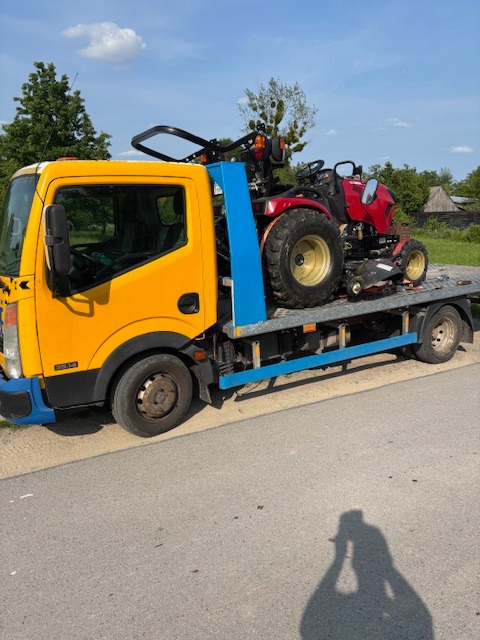 Żółta laweta przewozi czerwony traktor ogrodowy. Widoczne drzewa i błękitne niebo w tle. Transport sprzętu ogrodniczego w słoneczny dzień.