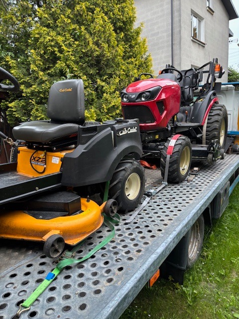 Czerwony traktor i żółta kosiarka Cub Cadet na przyczepie transportowej, zabezpieczone pasami, na tle budynku i zieleni.