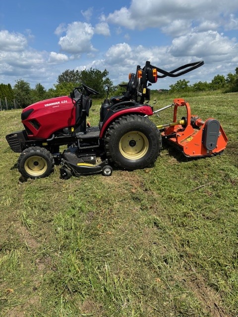 Czerwony traktor Yanmar kosi trawę na polu w słoneczny dzień. Świeżo skoszona trawa leży na ziemi. Widoczny fragment pola i drzewa w tle.