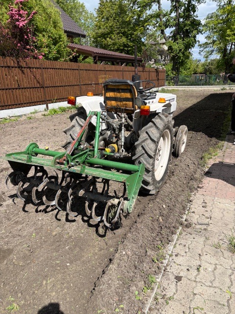 Mały traktor z glebogryzarką spulchnia ziemię na działce, przygotowując ją do siewu. Widok z tyłu na maszynę i świeżo obrobiony teren.