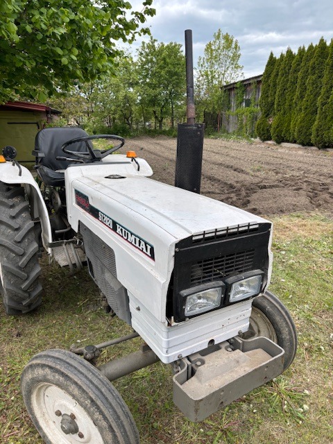 Biały traktor KUMAIAI ST2020 na tle przygotowanego pola i zieleni. Widoczny z bliska, z fragmentem drzewa po lewej stronie. Charakterystyczny czarny komin.