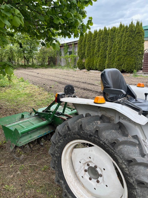 Biały traktor z zielonym glebogryzem na nierównym terenie, w tle rząd tuj i budynek gospodarczy. Widok zza koła, częściowo zasłaniającego maszynę.