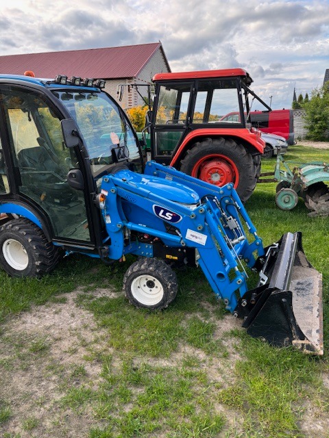 Niebieski traktor LS z łyżką na trawiastym terenie. W tle drugi traktor z czerwoną kabiną i budynek z czerwonym dachem. Sprzęt do robót ziemnych.