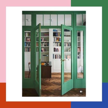 Otwarta, szklana, zielona rama drzwiowa prowadzi do domowej biblioteki z białymi regałami wypełnionymi książkami, biurkiem i parkietem ułożonym w jodełkę.