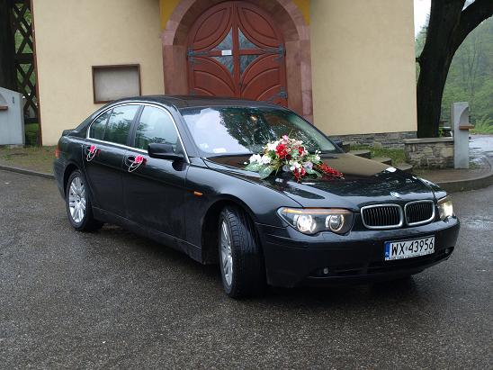 Czarna limuzyna BMW serii 7, udekorowana kwiatami na masce i kokardkami na klamkach, zaparkowana przed budynkiem z drewnianymi drzwiami.