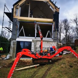 Expert Sp. z.o.o. - Montaż okna w surowym stanie budynku jednorodzinnego z użyciem czerwonego minikoparki z przyssawkami do szyb.