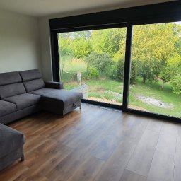 Expert Sp. z.o.o. - Przestronny salon z dużym, przesuwnym oknem z czarnymi ramami, widok na zielony ogród i jezioro, szara sofa narożna, drewniana podłoga.