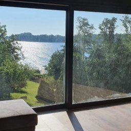 Expert Sp. z.o.o. - Duże przesuwne okno z czarnymi ramami, widok na jezioro i zieleń, fragment szarej sofy w rogu pomieszczenia.