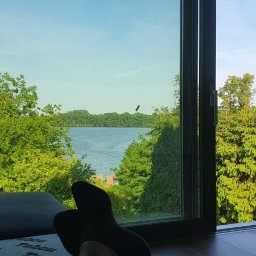 Expert Sp. z.o.o. - Widok z wnętrza przez otwarte, nowoczesne okno na jezioro otoczone zielenią, z fragmentem sofy i stóp w skarpetkach w kadrze.