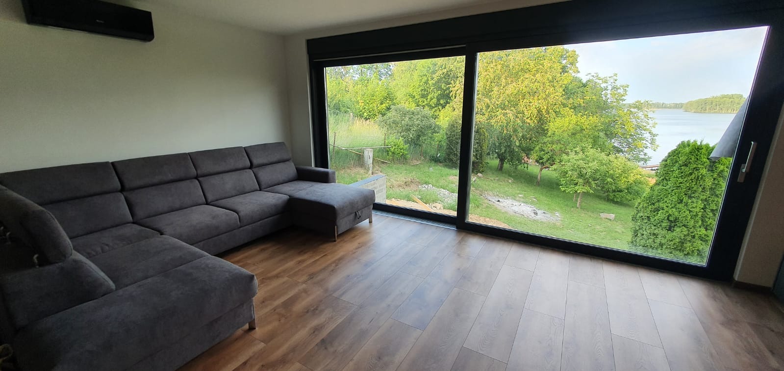 Przestronny salon z dużym, przesuwnym oknem z czarnymi ramami, widok na zielony ogród i jezioro, szara sofa narożna, drewniana podłoga.