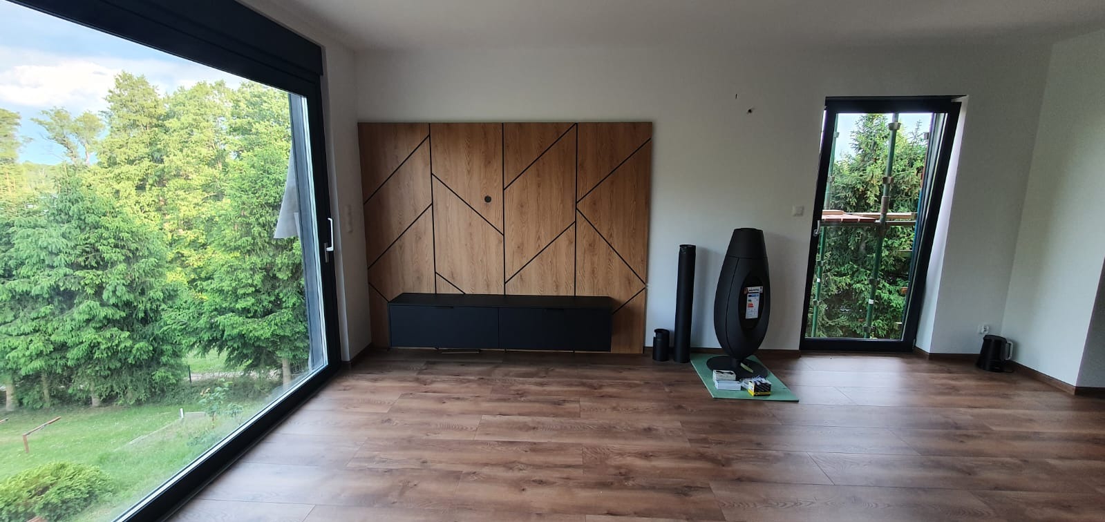 Wnętrze pokoju z dużym oknem z czarną ramą, widok na zieleń za oknem, drewniana ściana dekoracyjna z czarnymi liniami, czarna szafka RTV i czarny piec wolnostojący na podłodze imitującej drewno.
