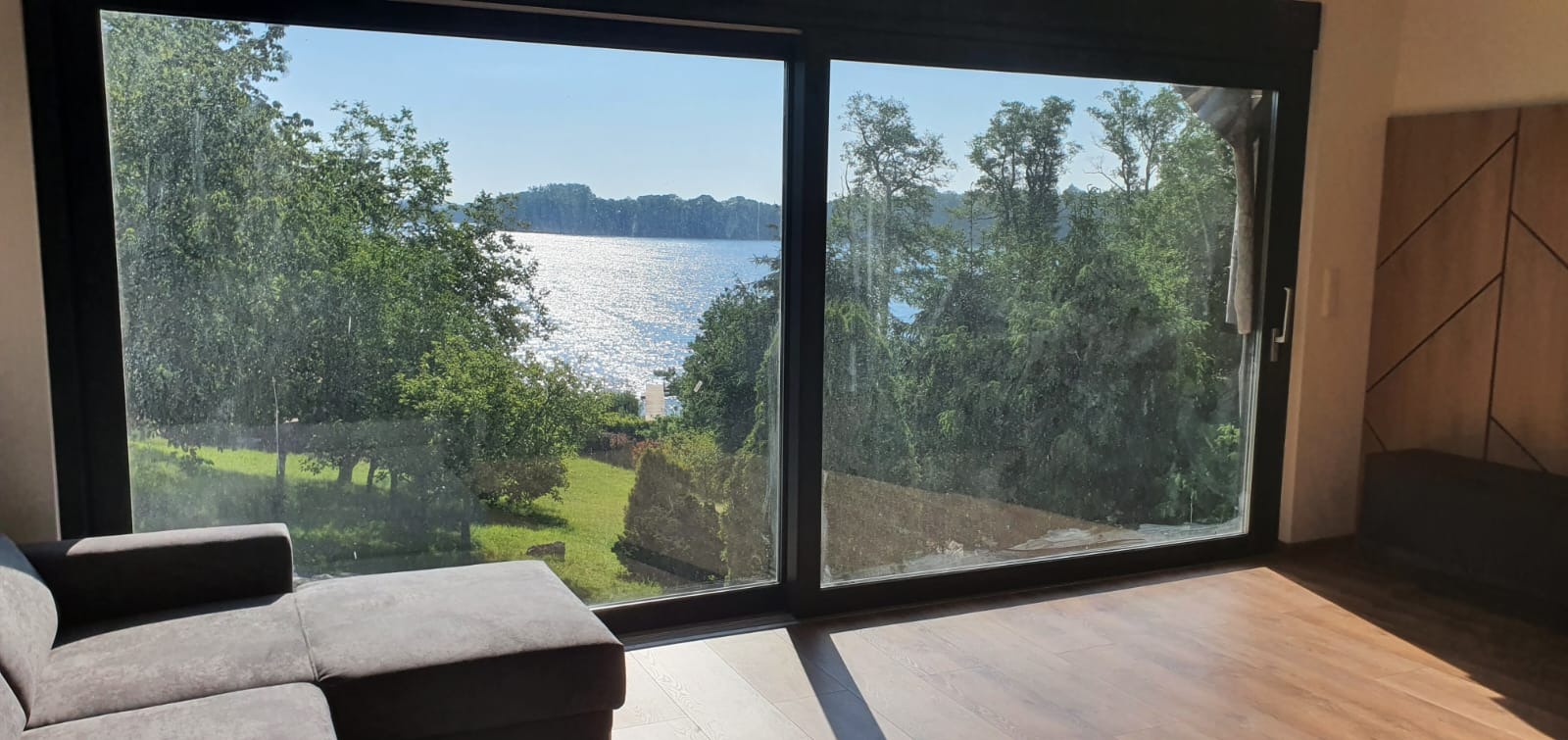 Duże przesuwne okno z czarnymi ramami, widok na jezioro i zieleń, fragment szarej sofy w rogu pomieszczenia.