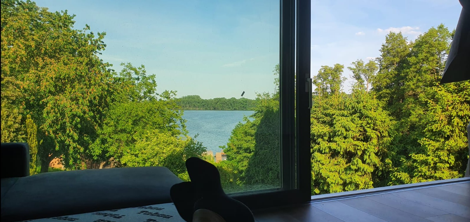 Widok z wnętrza przez otwarte, nowoczesne okno na jezioro otoczone zielenią, z fragmentem sofy i stóp w skarpetkach w kadrze.