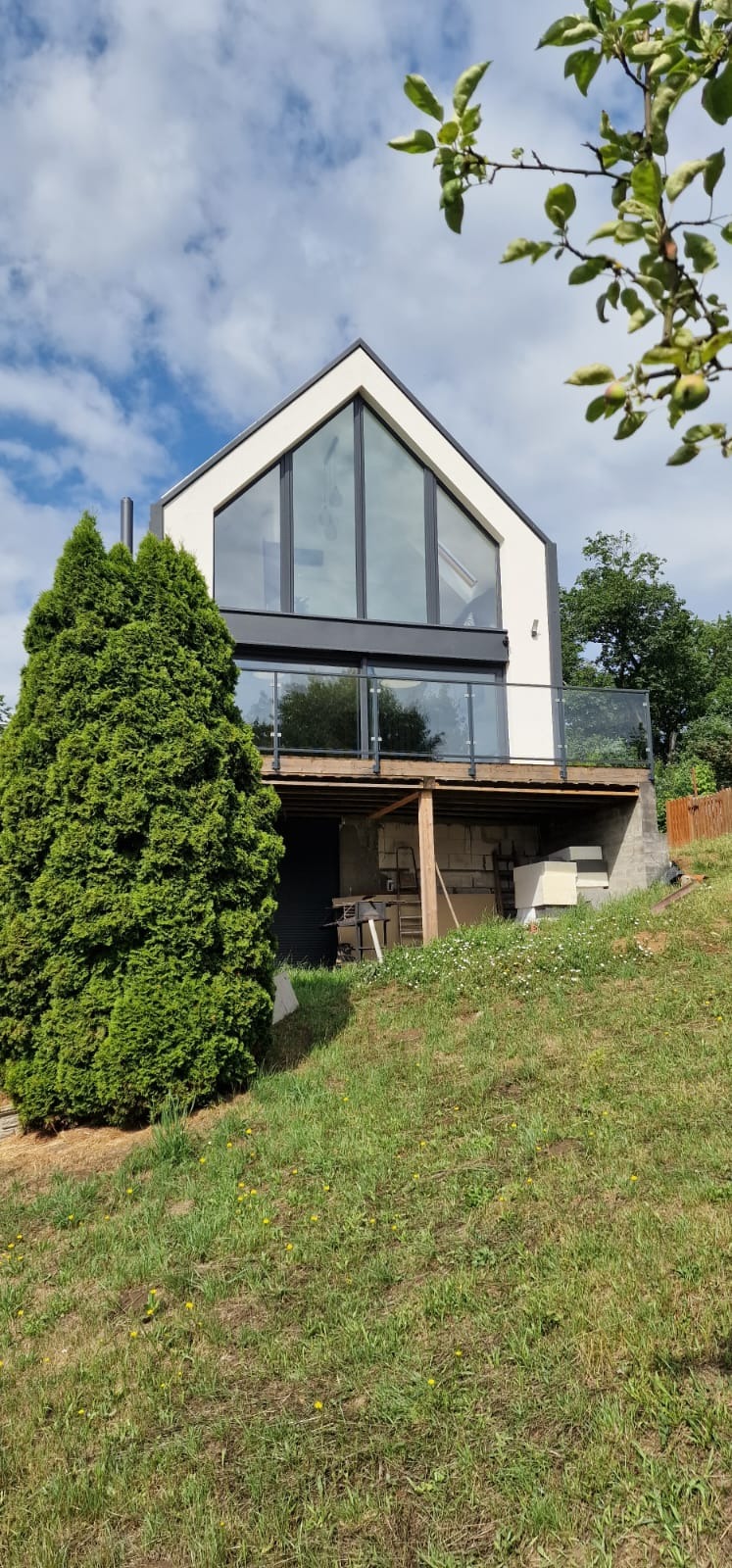 Nowoczesny dom z dużymi oknami aluminiowymi i szklaną balustradą na tarasie, widok z dołu na tle zieleni i błękitnego nieba z chmurami.