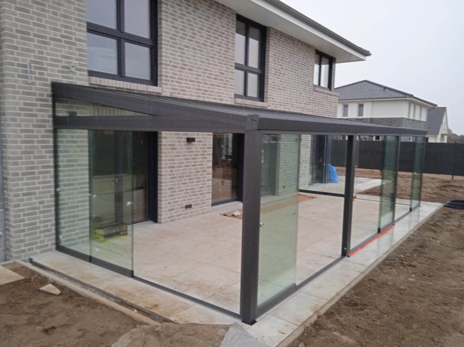 Szklana zabudowa tarasu z przesuwnymi drzwiami i grafitową ramą, przylegająca do szarej ściany z cegły klinkierowej; widoczne okna w tej samej tonacji oraz fragment posesji.