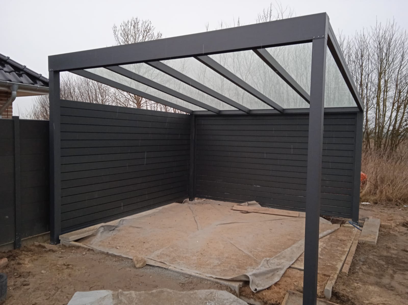 Nowoczesna, szara pergola z przezroczystym zadaszeniem i panelami bocznymi z desek kompozytowych, widoczna na placu budowy z rozłożoną plandeką ochronną.