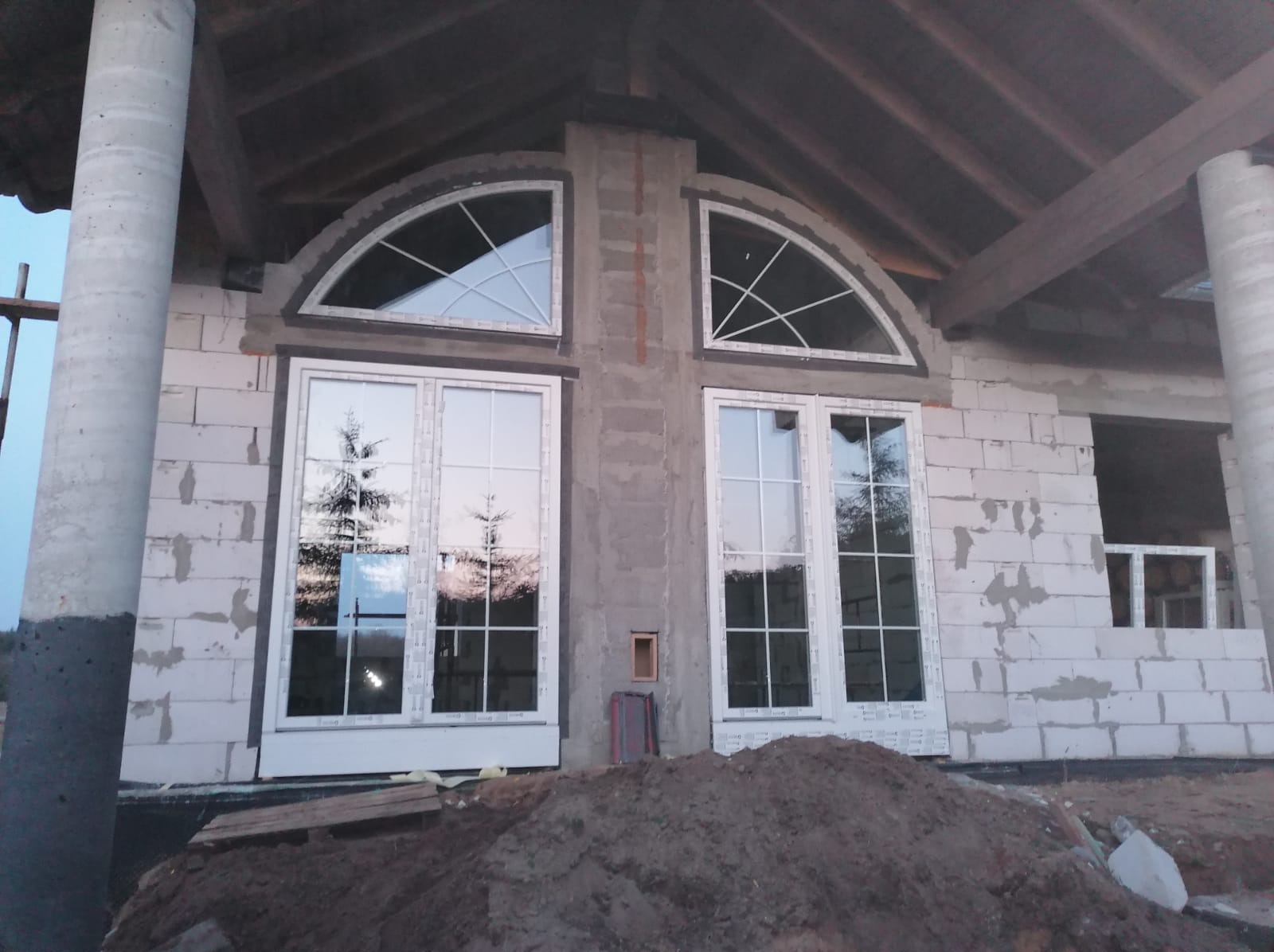Montaż białych okien z łukowym nadprożem w surowym stanie budynku, widoczne bloczki betonowe i ziemia.
