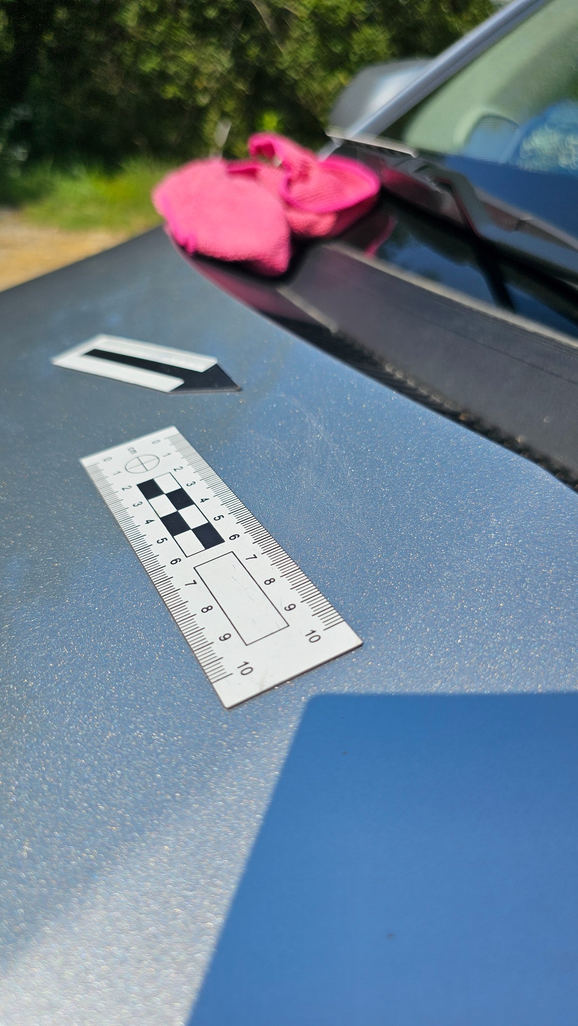 Błyszcząca powierzchnia karoserii samochodu z widocznymi rysami, obok leży biała miarka z podziałką centymetrową i znacznik, w tle różowa ściereczka