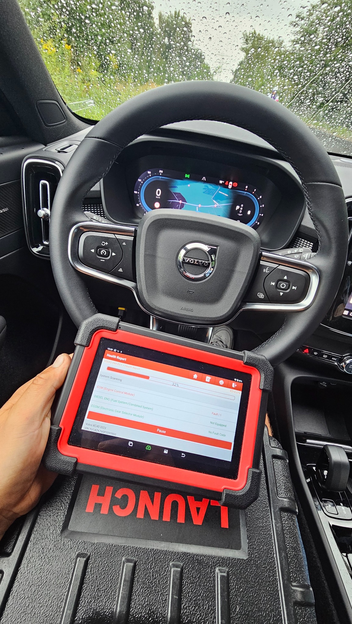 Wnętrze samochodu Volvo XC40 z tabletem diagnostycznym marki Launch na desce rozdzielczej, wyświetlającym raport o stanie systemów pojazdu podczas skanowania. Na przedniej szybie widoczne krople...