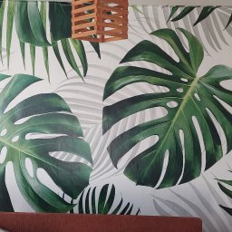 Team-Lewy - Ściana z fototapetą w motywie liści monstery i palm, drewniana lampa wisząca, widoczne niedociągnięcia wykończeniowe na krawędzi ściany.