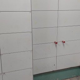 Team-Lewy - Ściana w trakcie układania płytek imitujących panele, widoczne plastikowe krzyżyki dystansowe, otwory na instalacje i zabrudzenia po kleju.