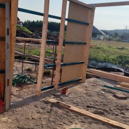 Team-Lewy - Konstrukcja szkieletowa budynku w trakcie budowy, widoczne drewniane elementy, płyta OSB, poziomica, narzędzia i piasek na podłożu.