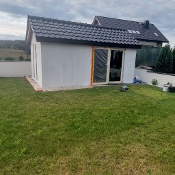 Team-Lewy - Nowo otynkowany budynek gospodarczy z ciemnym dachem, folią zabezpieczoną stolarką okienną i drzwiową, stojący na trawniku przed domem jednorodzinnym w pochmurny dzień.