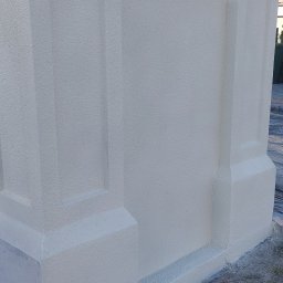 Team-Lewy - Fragment ocieplonej elewacji budynku z wyraźną strukturą baranka i dekoracyjnymi elementami architektonicznymi, widoczne ślady po pracach wykończeniowych u podstawy.