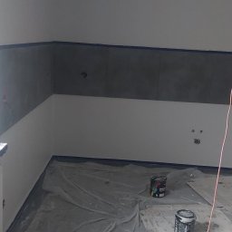 Team-Lewy - Przygotowane do malowania wnętrze: zabezpieczona folią podłoga, częściowo pomalowane ściany z taśmą malarską, widoczne puszki z farbą.