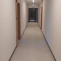 Team-Lewy - Długi korytarz w nowym budynku z białymi ścianami, jasną podłogą i drewnianymi drzwiami po bokach, zakończony czarnymi drzwiami ewakuacyjnymi.