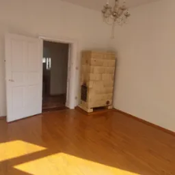 Pokój z drewnianą podłogą, otwartymi białymi drzwiami, kaflowym piecem i kryształowym żyrandolem