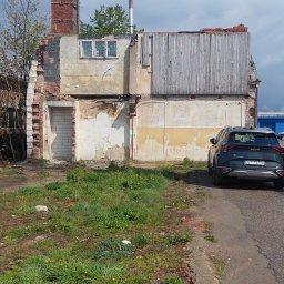 Team-Lewy - Zrujnowany budynek z częściowo zamurowanym oknem i drzwiami, obok zaparkowany czarny samochód.