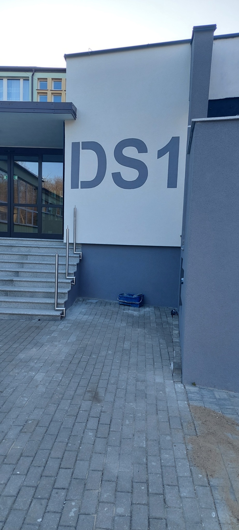 Ocieplona elewacja budynku z szarym napisem 'DS1', wejście z granitowymi schodami i metalowymi barierkami, oraz niebieskie wiadro na kostce brukowej.