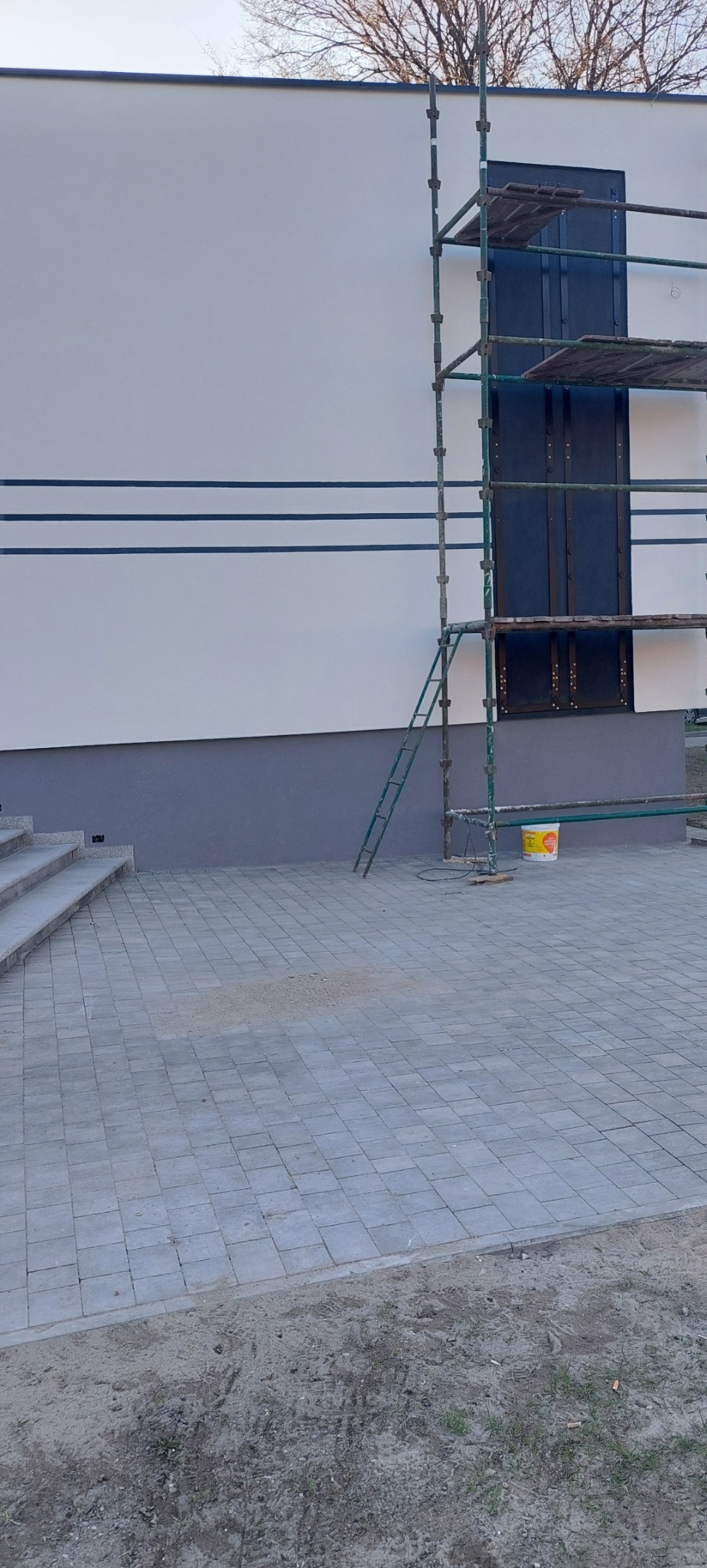 Ściana budynku po malowaniu na biało, z szarym cokołem i trzema poziomymi, ciemnymi pasami. Przy ścianie stoi metalowe rusztowanie, a obok niego wiadro z farbą. Podłoże wyłożone szarą kostką brukową.