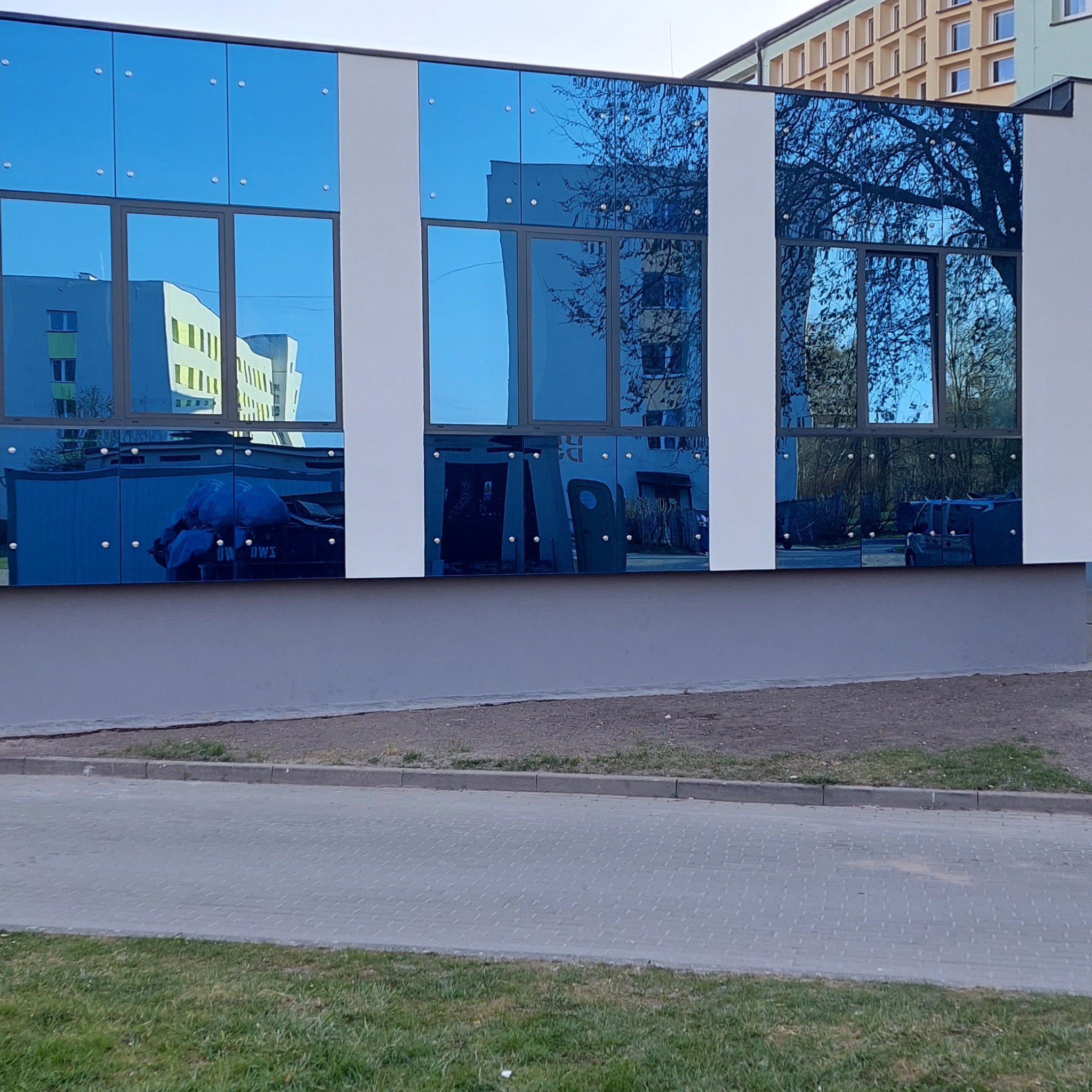 Nowoczesna fasada budynku z niebieskimi, lustrzanymi panelami, odbijającymi otoczenie, w tym inne budynki i drzewa.