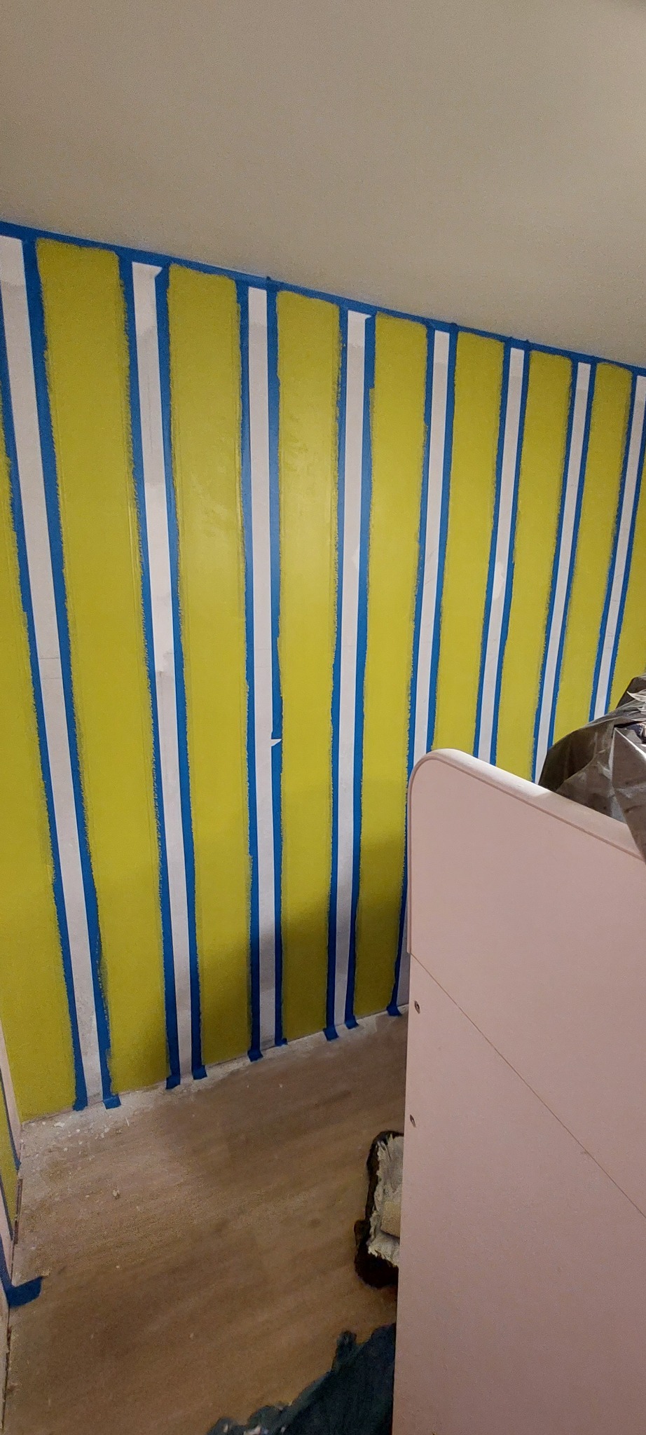 Świeżo pomalowana ściana w pionowe pasy w kolorze żółtym i białym, oddzielone niebieską taśmą malarską, widoczny fragment podłogi i mebla w rogu pomieszczenia.