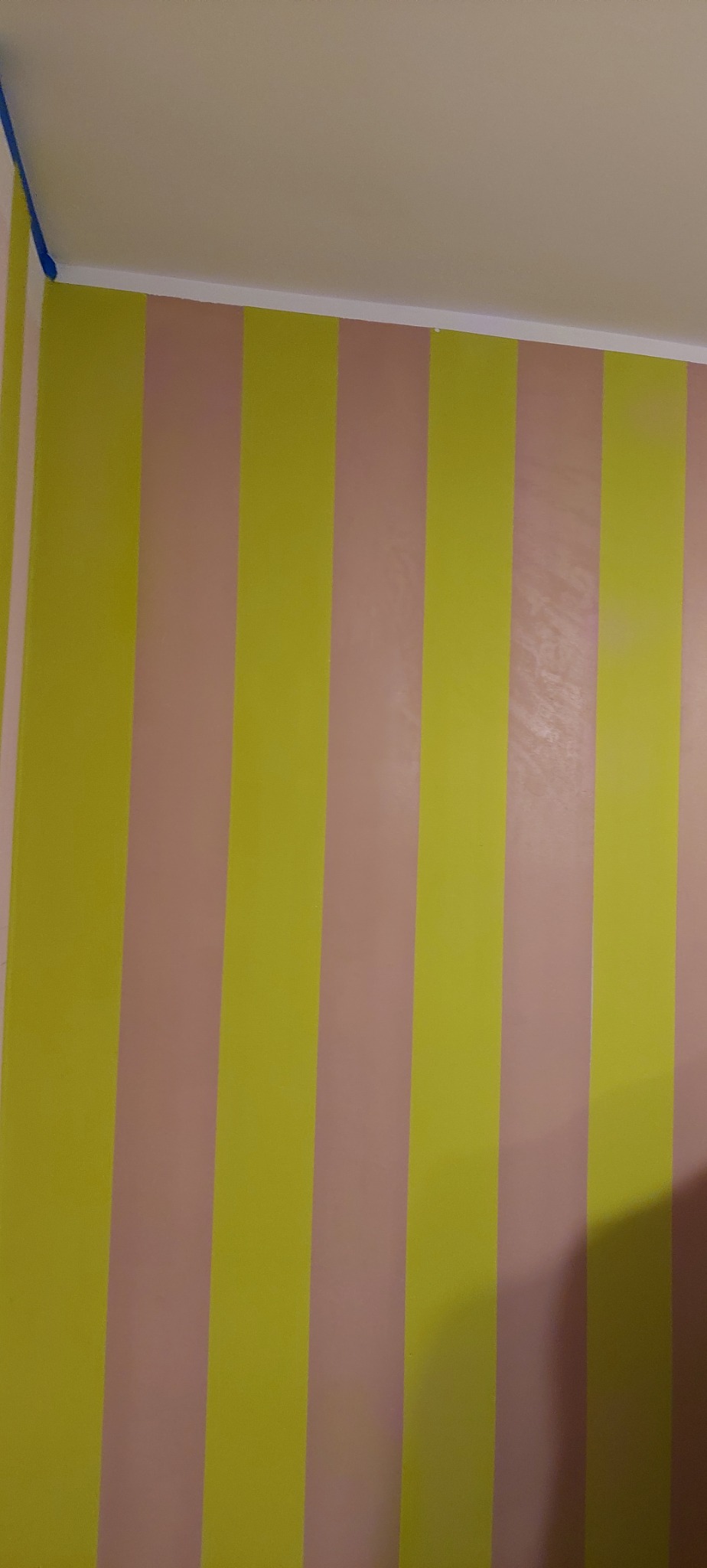 Pomalowana ściana w pionowe pasy w odcieniach żółci i pudrowego różu, widoczne niebieskie taśmy malarskie przy suficie.