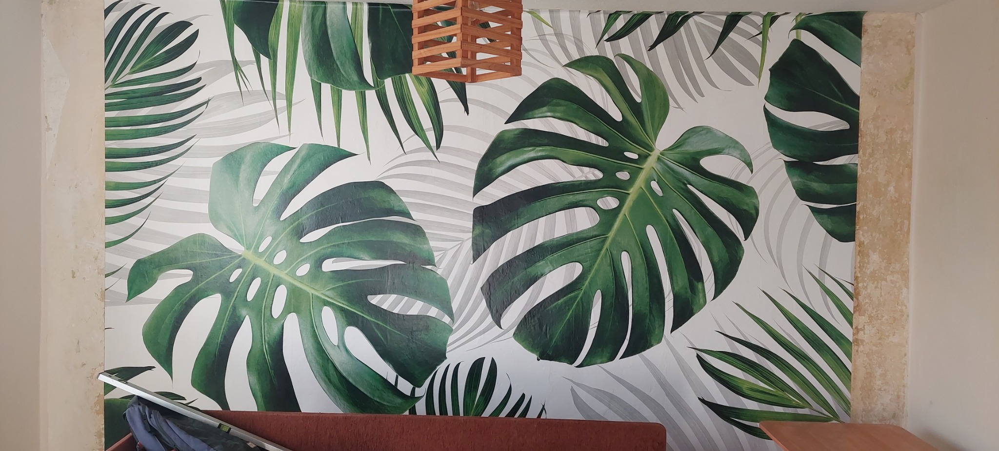 Ściana z fototapetą w motywie liści monstery i palm, drewniana lampa wisząca, widoczne niedociągnięcia wykończeniowe na krawędzi ściany.