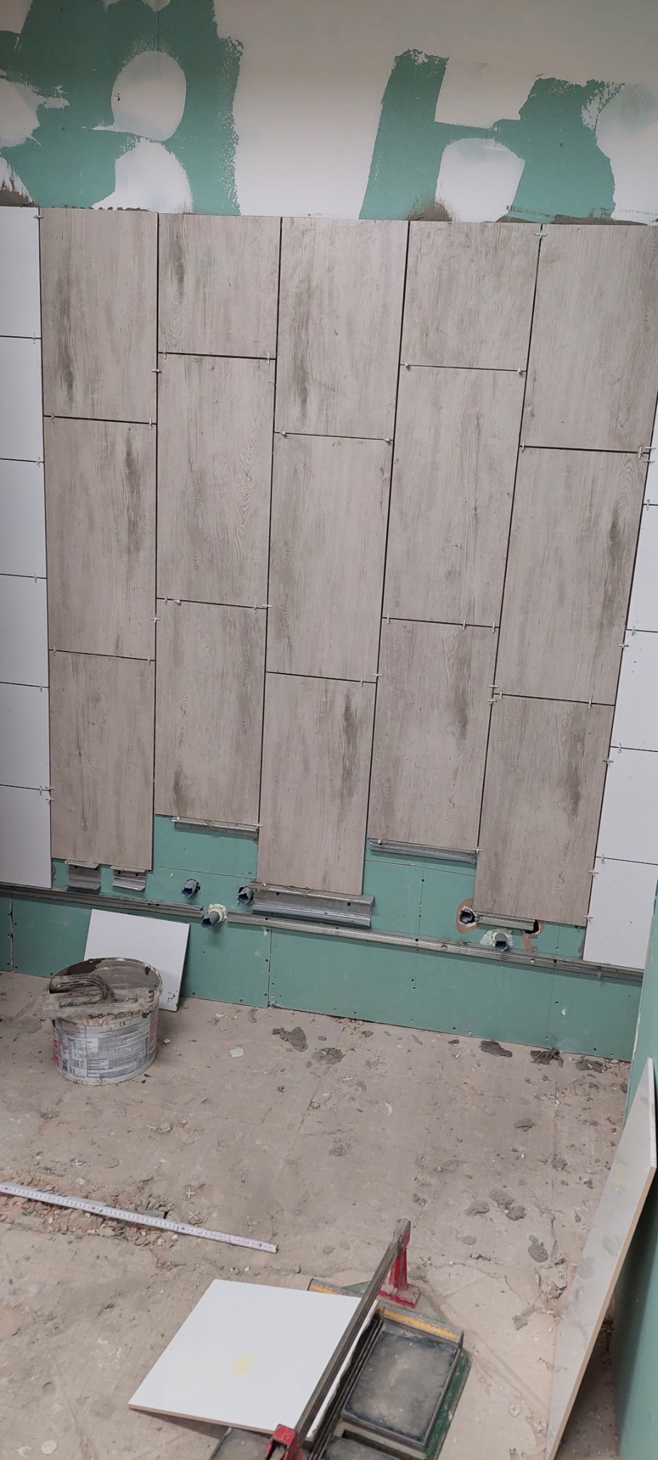 Montaż płytek imitujących drewno na ścianie z płyt gipsowo-kartonowych w trakcie remontu, widoczne listwy montażowe, wiadro z zaprawą i gilotyna do cięcia płytek.