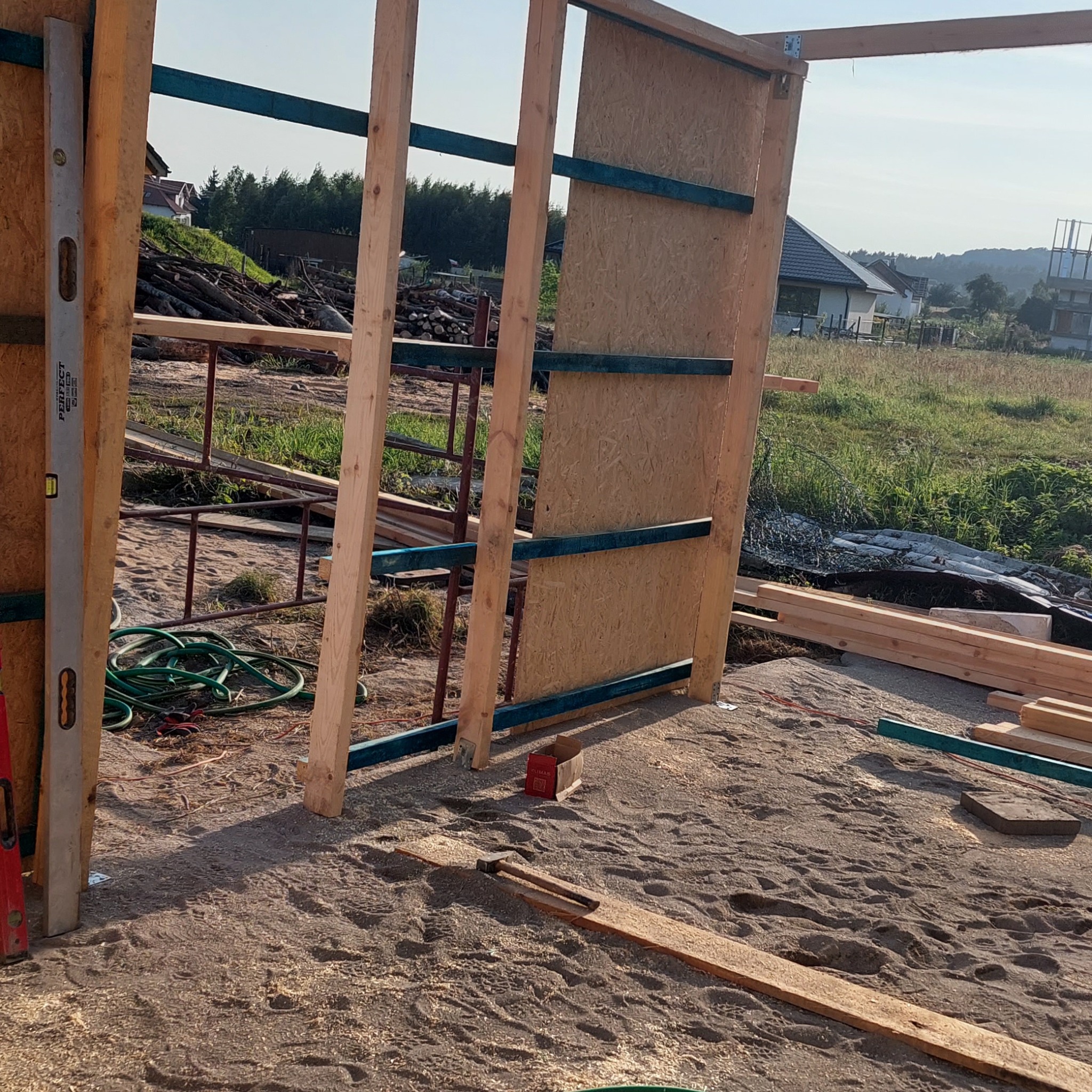 Konstrukcja szkieletowa budynku w trakcie budowy, widoczne drewniane elementy, płyta OSB, poziomica, narzędzia i piasek na podłożu.