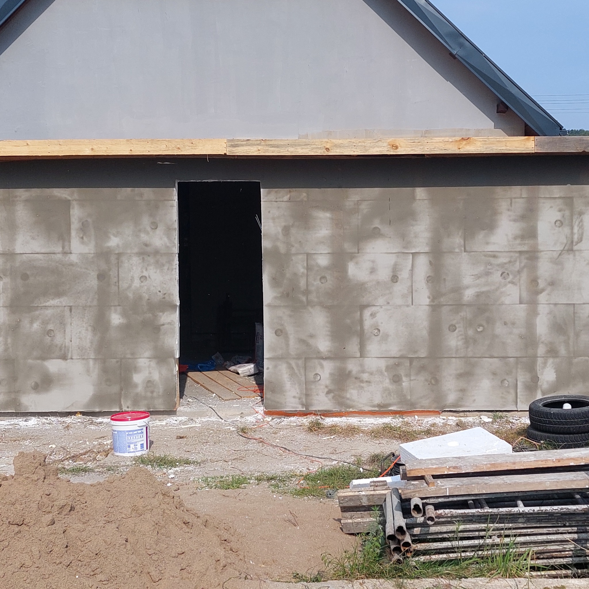 Ściana budynku w trakcie ocieplania z widocznymi panelami izolacyjnymi, otwartym otworem drzwiowym, stertą piasku, puszką farby i stosem metalowych rur na pierwszym planie.