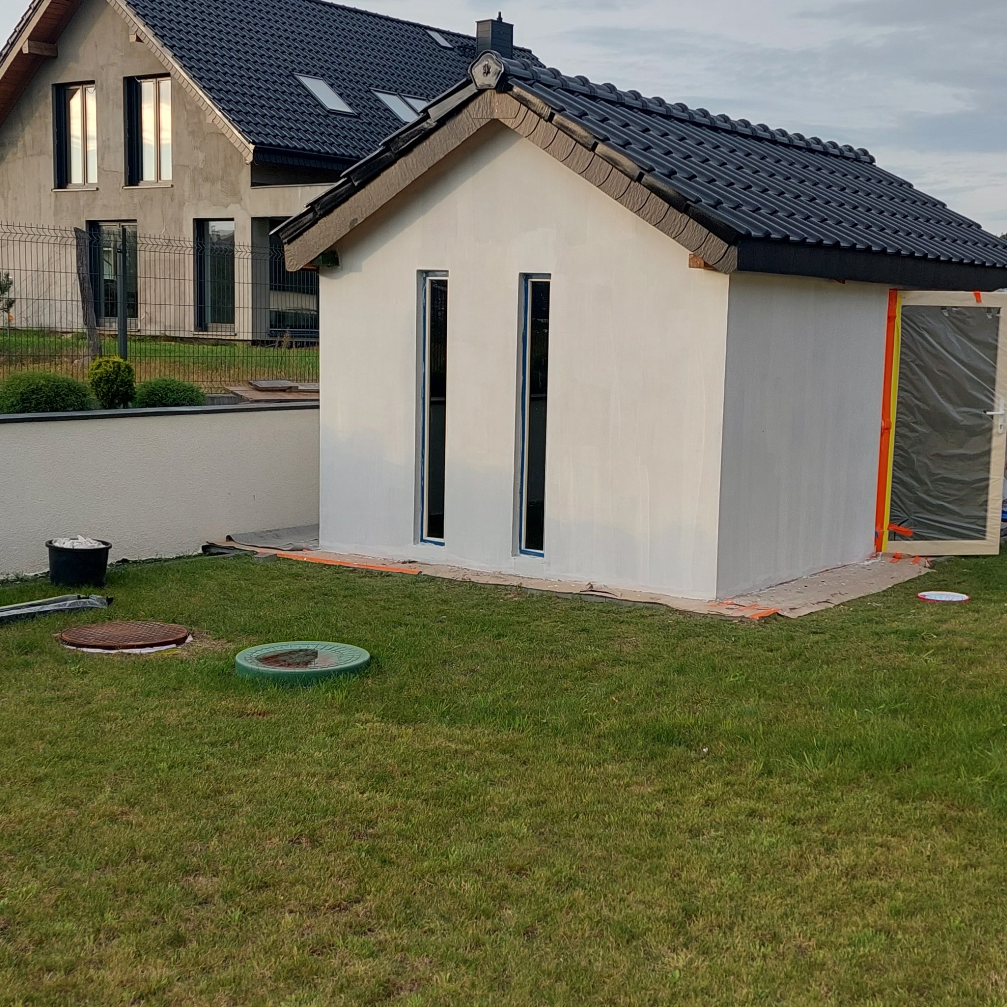 Ocieplony budynek gospodarczy z dwoma pionowymi oknami, świeżo pomalowany na biało, z zabezpieczonym wejściem folią i taśmą malarską, widoczne manewry na trawie i fragment domu w tle.