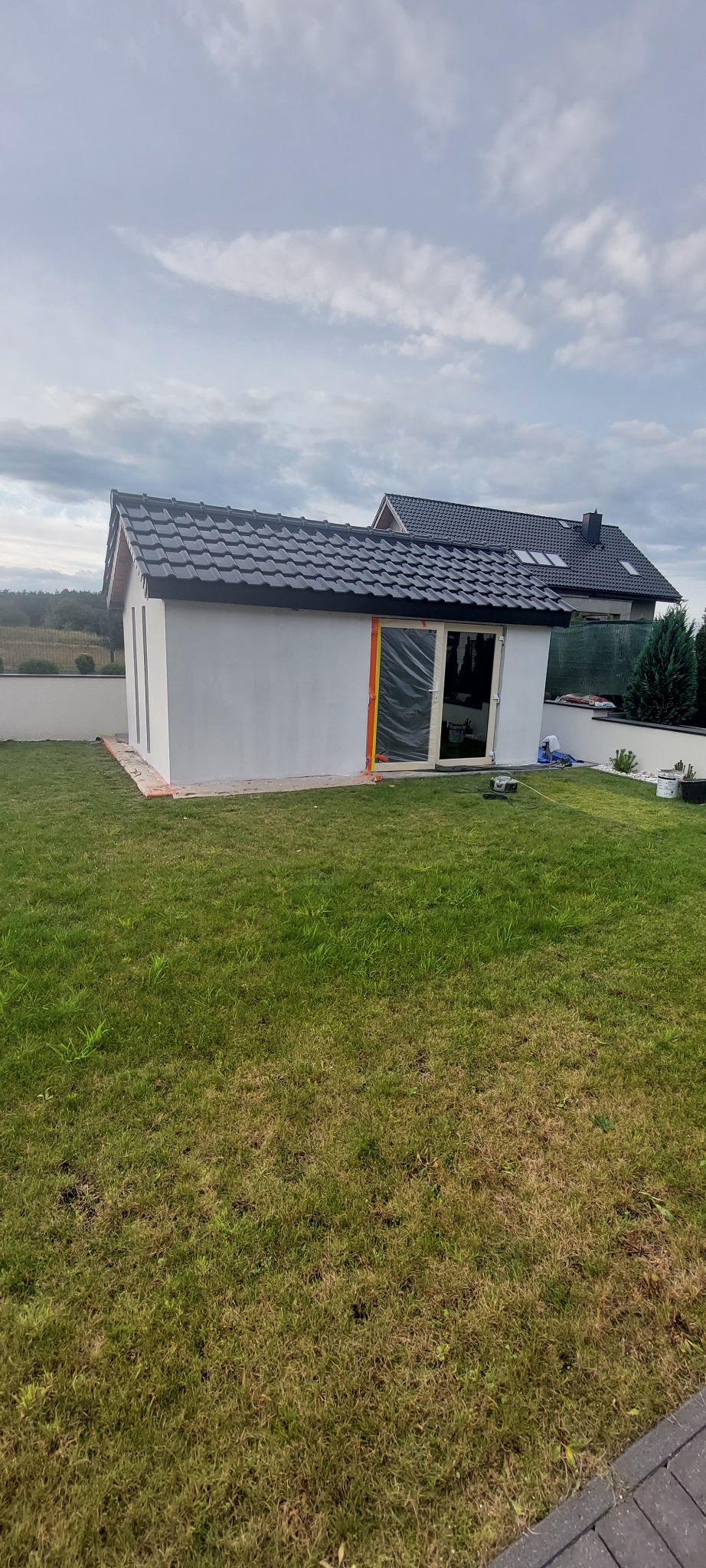 Nowo otynkowany budynek gospodarczy z ciemnym dachem, folią zabezpieczoną stolarką okienną i drzwiową, stojący na trawniku przed domem jednorodzinnym w pochmurny dzień.