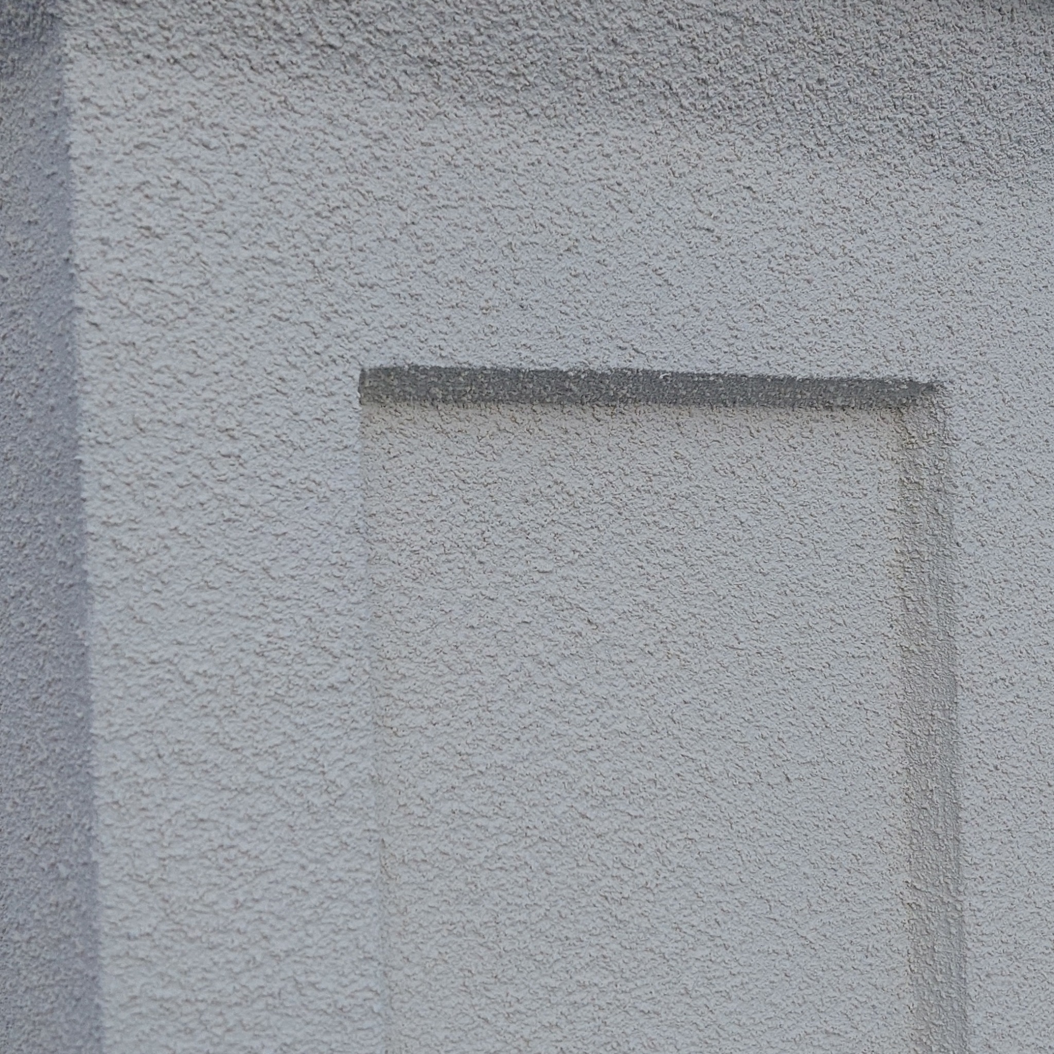 Fragment szarej ściany zewnętrznej z fakturą baranka i wklęsłym, prostokątnym elementem dekoracyjnym.