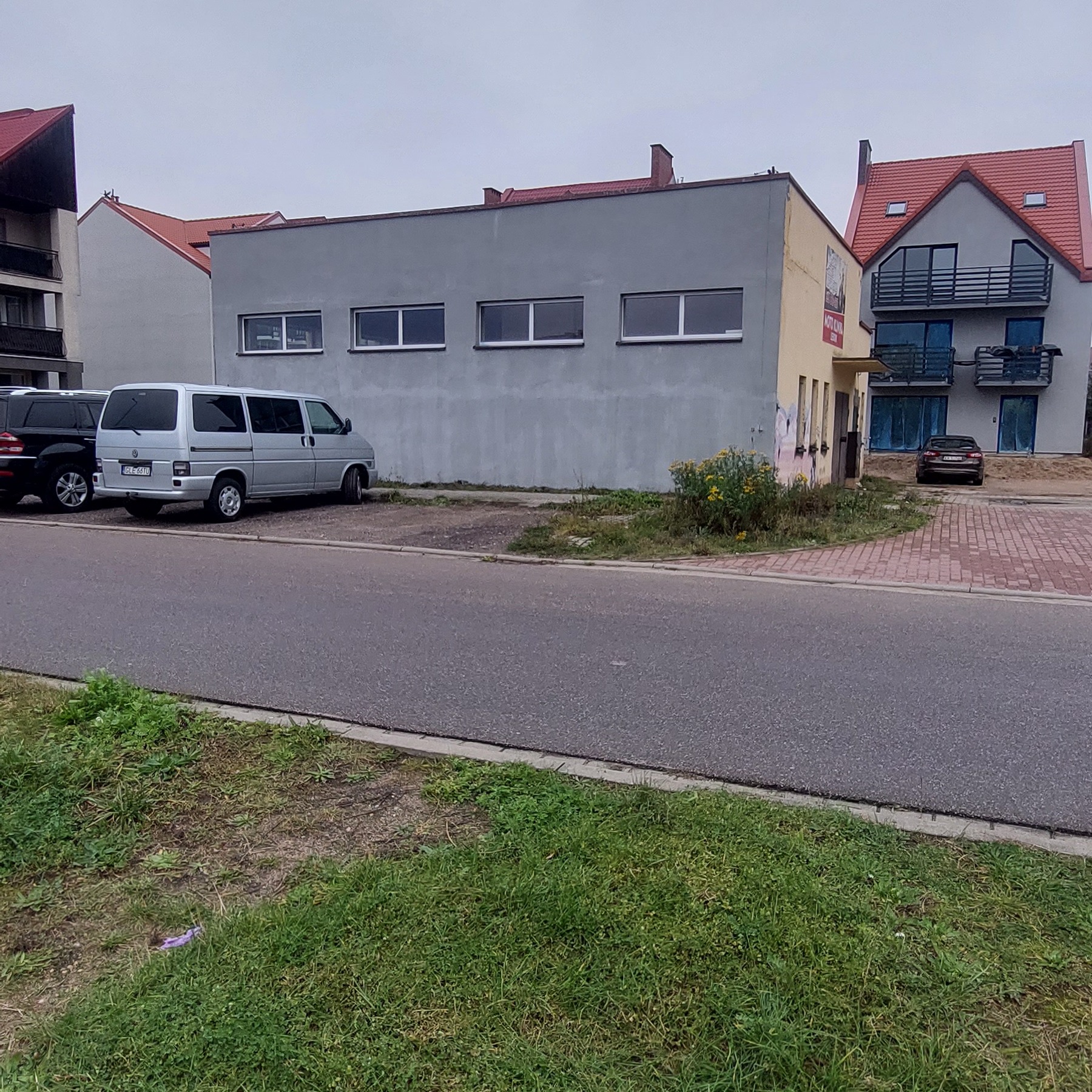 Szary budynek z trzema oknami i zaparkowanymi samochodami na tle innych budynków mieszkalnych.