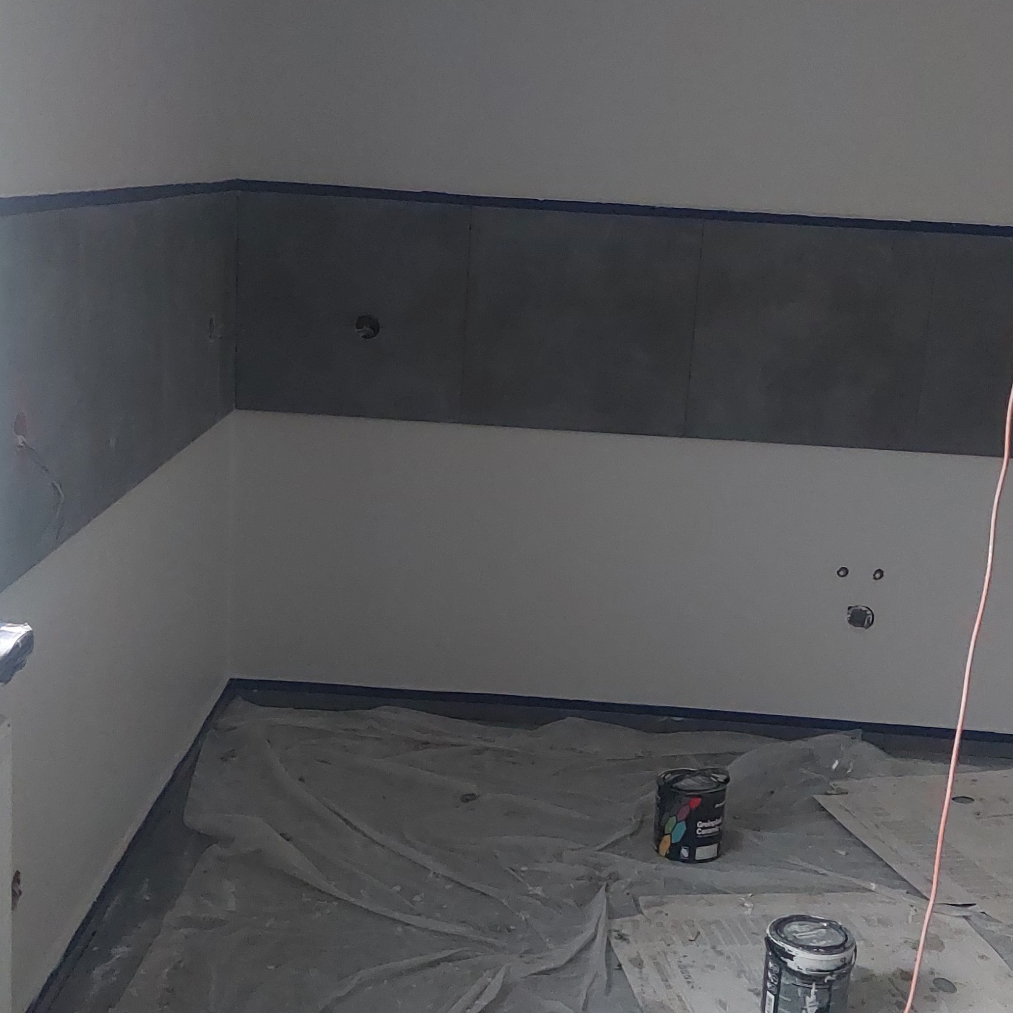 Przygotowane do malowania wnętrze: zabezpieczona folią podłoga, częściowo pomalowane ściany z taśmą malarską, widoczne puszki z farbą.