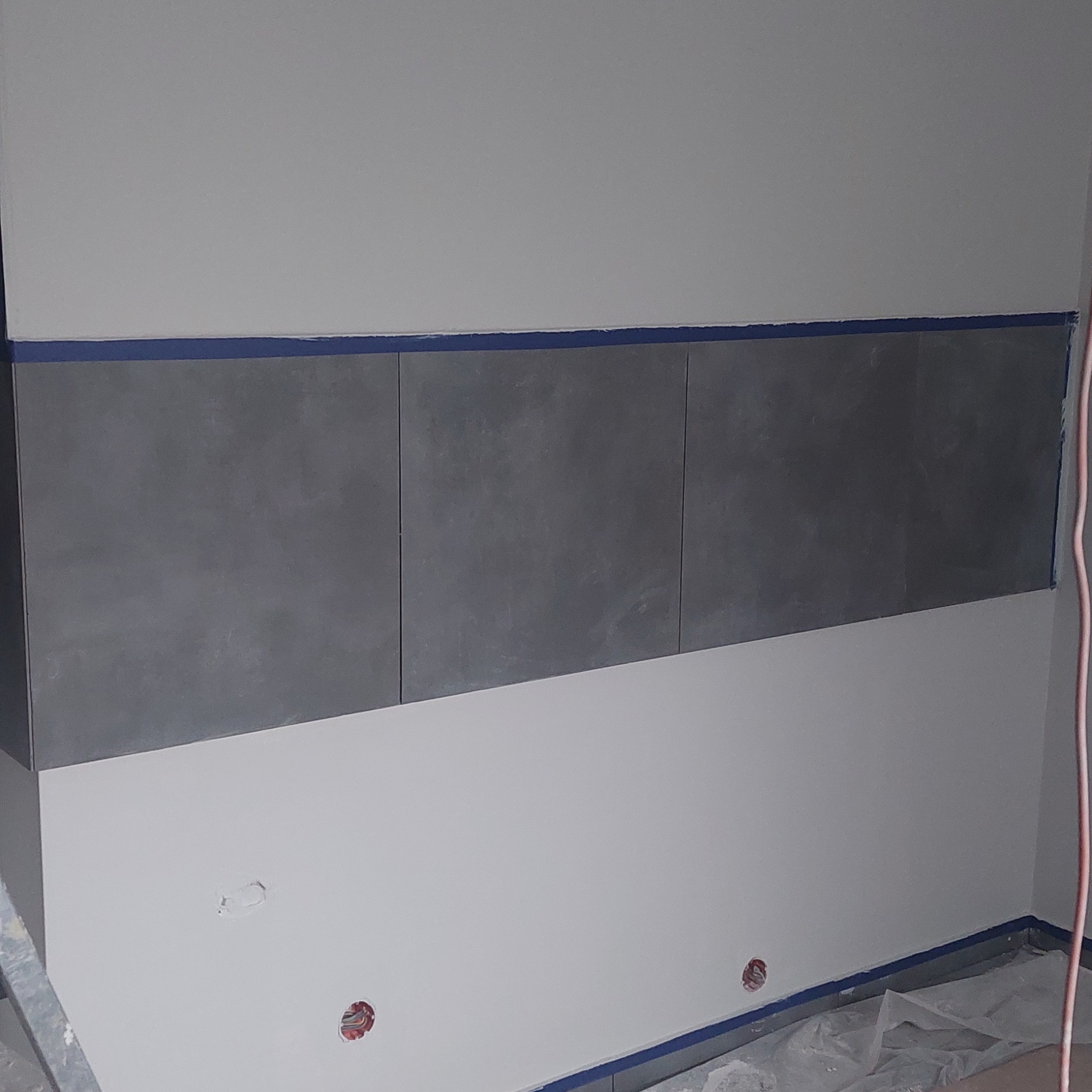 Ściana w trakcie remontu z trzema rzędami szarych płytek zabezpieczonych niebieską taśmą malarską, widoczne puszki elektryczne i folia ochronna na podłodze.